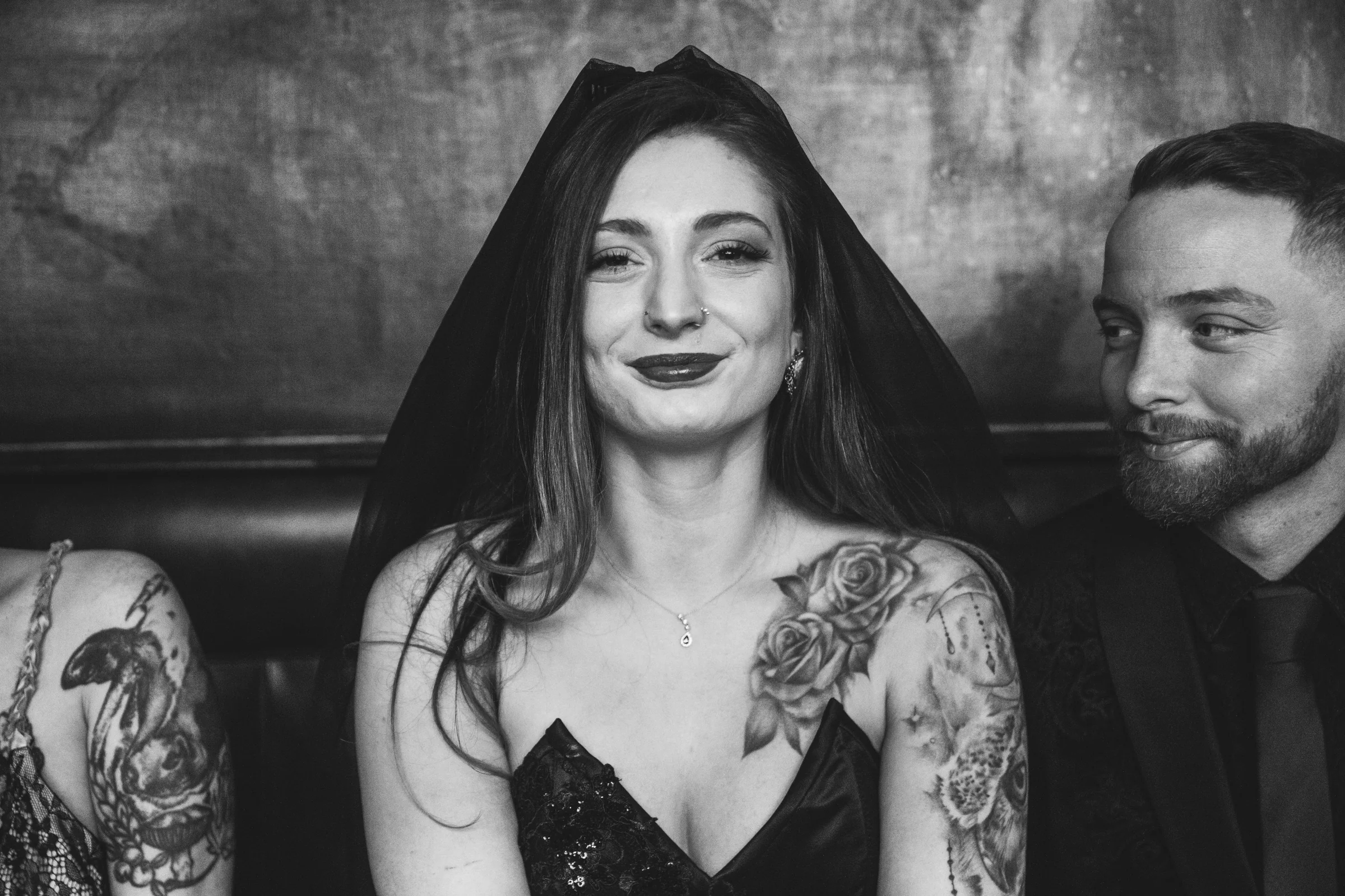 A tattooed bride in a black veil smiling in a black-and-white portrait